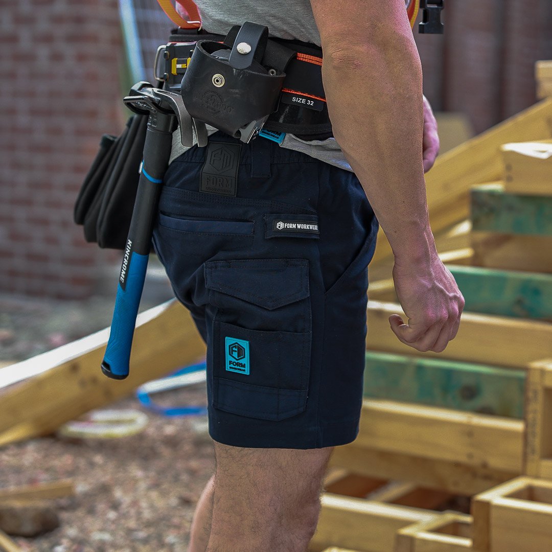 Tradesman wearing Form Workwear navy cargo shorts with a tool belt and hammer at a construction site.