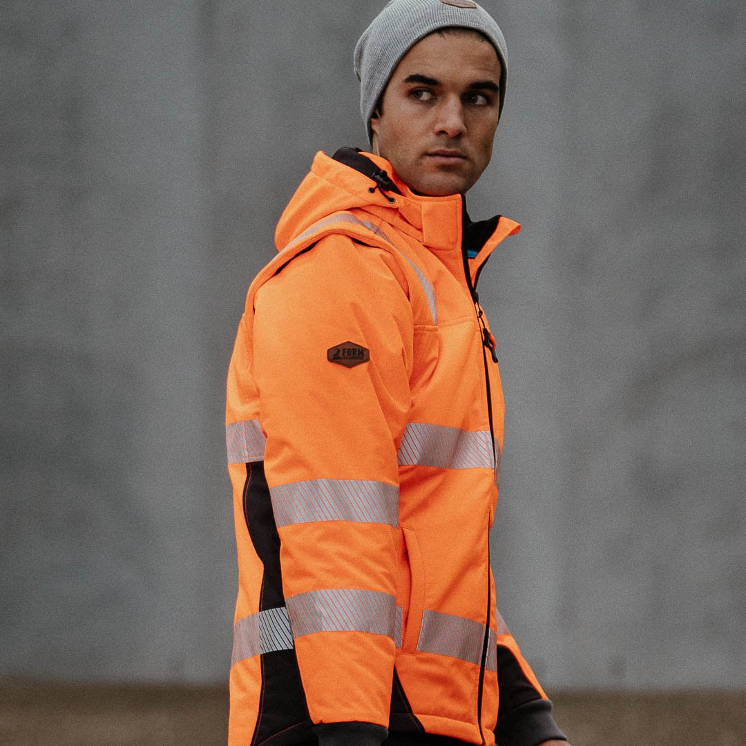 Young tradesman wearing high-visibility orange work jacket with reflective stripes, looking off to the side against a neutral background