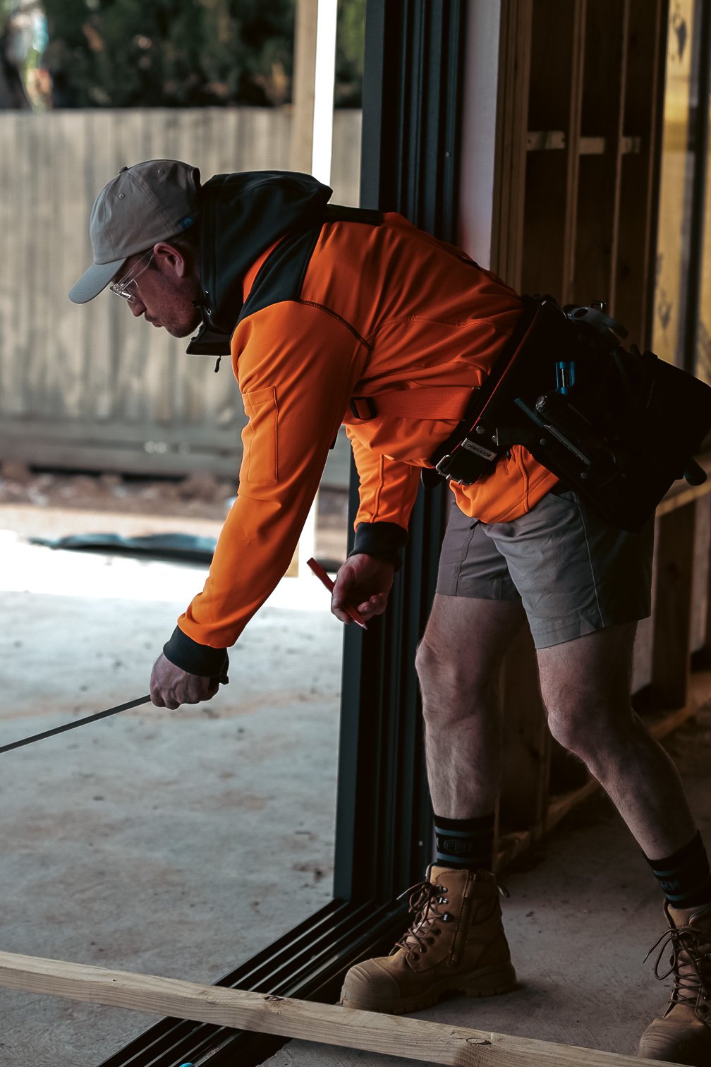 Construction worker measuring with a tape while wearing a hi-vis orange jacket, utility belt, and beige cap inside a building under construction