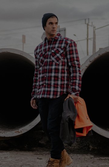 Tradesman wearing red flannel shirt, black pants and boots holding hi-vis gear in front of concrete pipes on jobsite