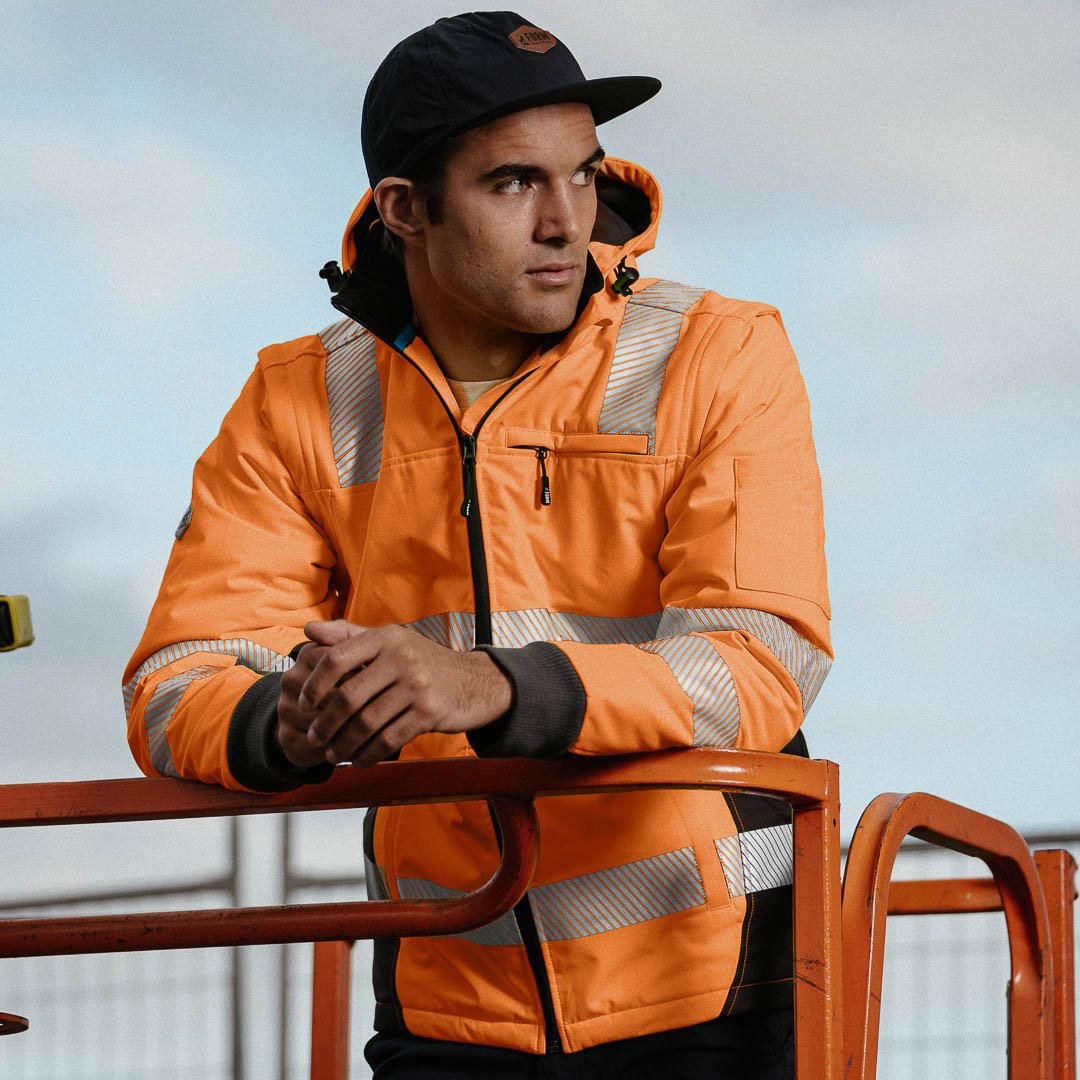 Worker in a hi-vis orange jacket with reflective tape and black cap, leaning on construction equipment outdoors
